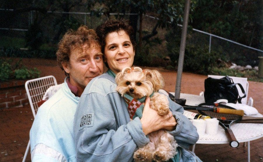 Gilda is sitting on Gene’s lap beside the tennis court as she holds their dog up to her chest.
