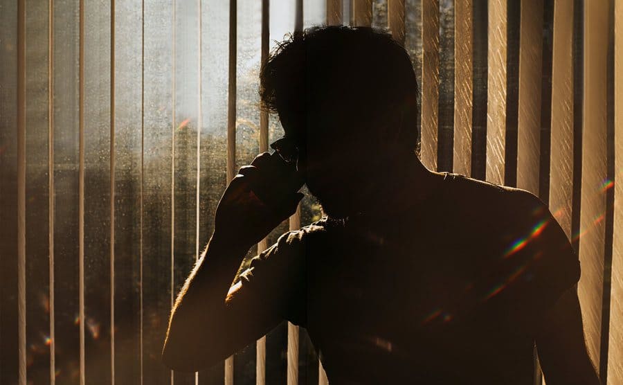 A silhouette of an unknown man on a phone standing against window blinds.