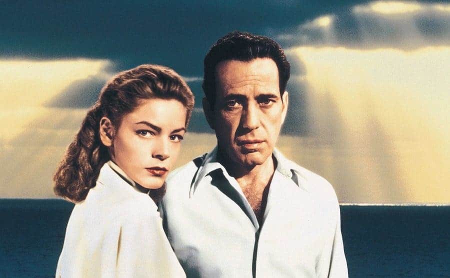 Lauren Bacall and Humphrey Bogart stand under the sunset in a film promo shot.