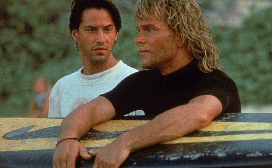 Reeves stands on a beach as Swayze holds a surfboard during the filming of 'Point Break'. 