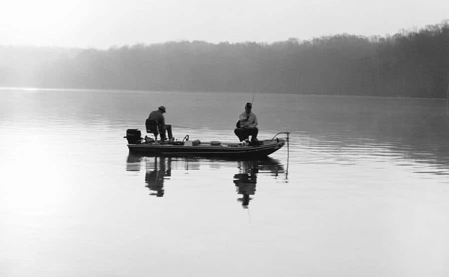 Two men are fishing, sitting in a small boat at dawn.