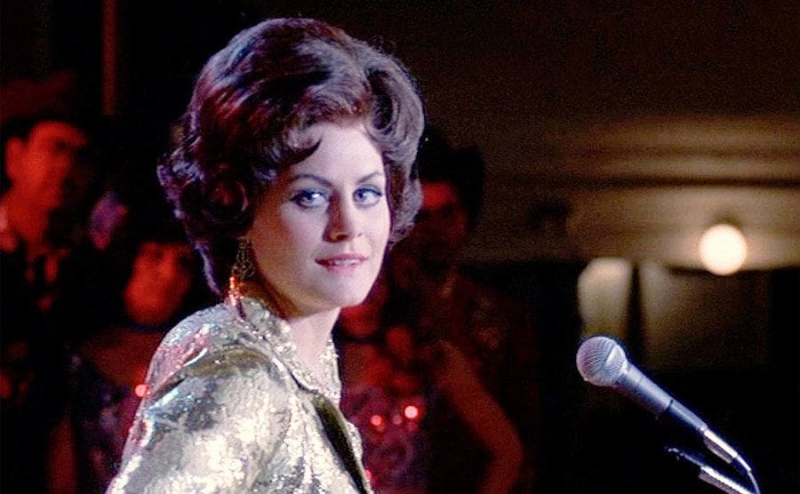 D’Angelo as Patsy Cline is standing on stage behind the microphone.