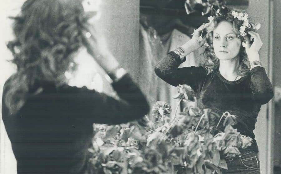 Beverly D’Angelo looks at her reflection in the mirror wearing a crown of flowers.