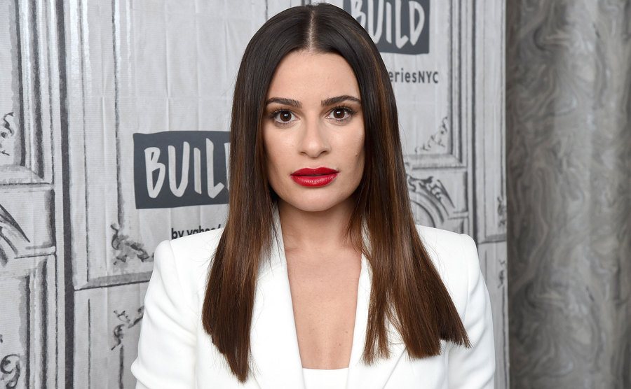 Lea Michele staring into the camera sporting red lipstick and a white suit.