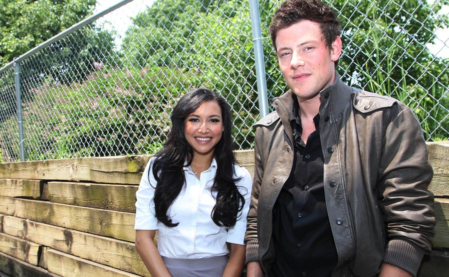 Naya Rivera and Cory Monteith stand beside each other near a fence.