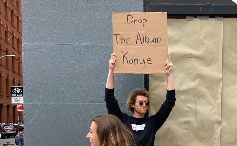 A man is holding a sign that reads ‘’Drop the Album Kanye.’’