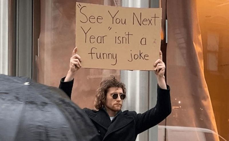 A man is holding a sign that reads ‘’See You Next Year isn’t a Funny Joke.’’