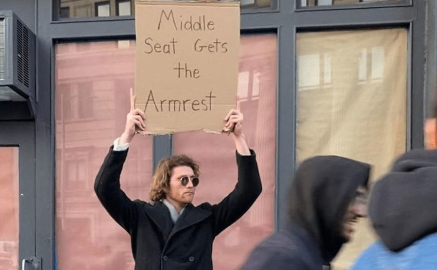 A man is holding a sign that reads ‘’Middle Seat Gets the Armrest.’’