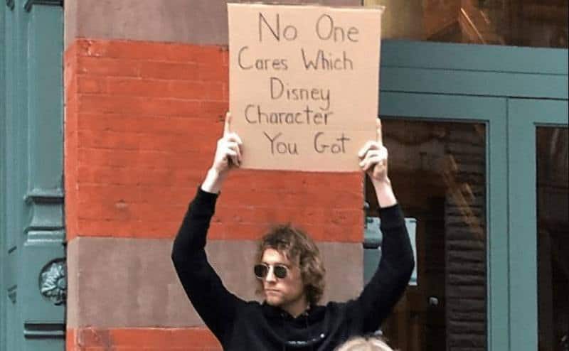 A man is holding a sign that reads ‘’No One Cares Which Disney Character You Got.’’