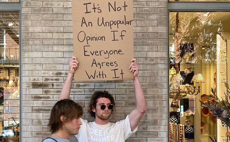 A man is holding a sign that reads ‘’It’s Not an Unpopular Opinion if Everyone Agrees with it.’’