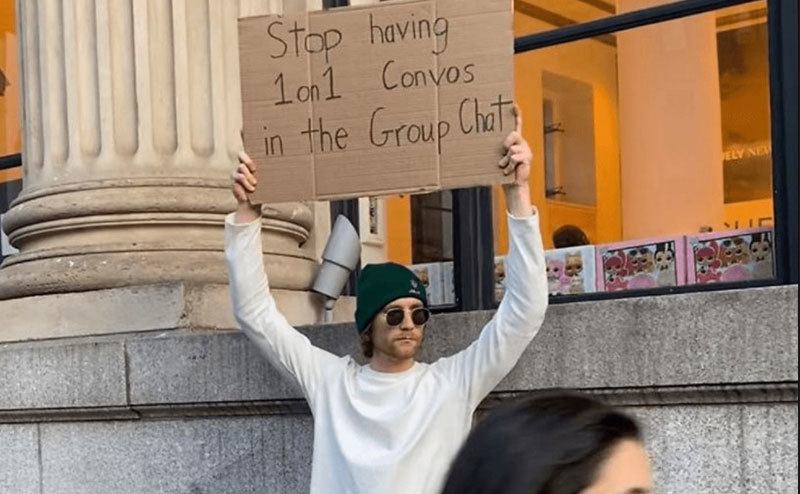 A man is holding a sign that reads ‘’Stop Having 1 on 1 Convos in the Group Chat.’’
