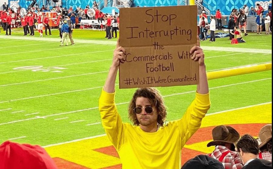 A man is holding a sign that reads ‘’Stop Interrupting the Commercials with Football.’’