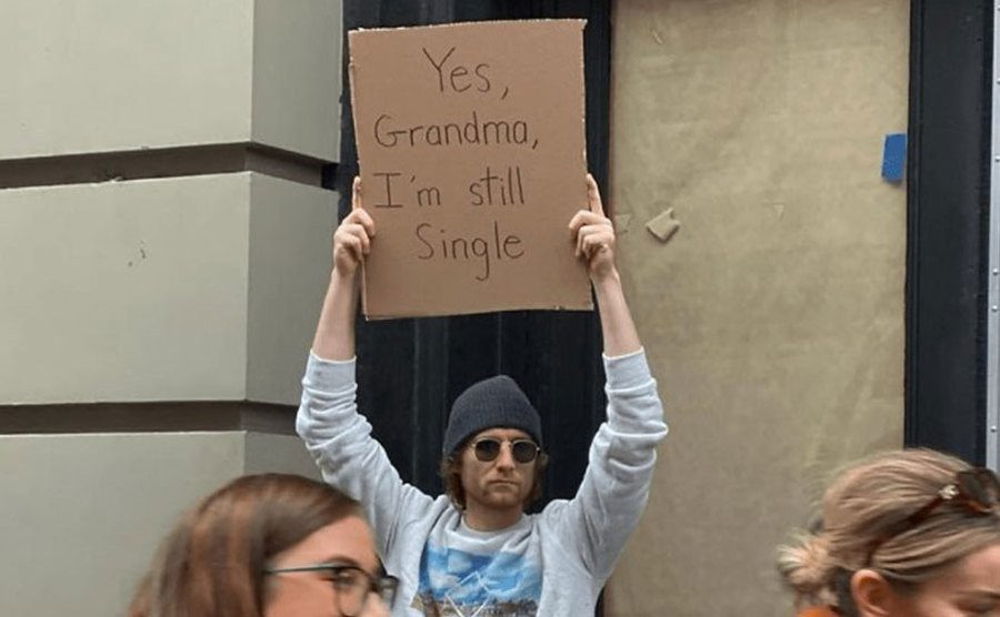 A man is holding a sign that reads ‘’Yes, Grandma, I’m Still Single.’’
