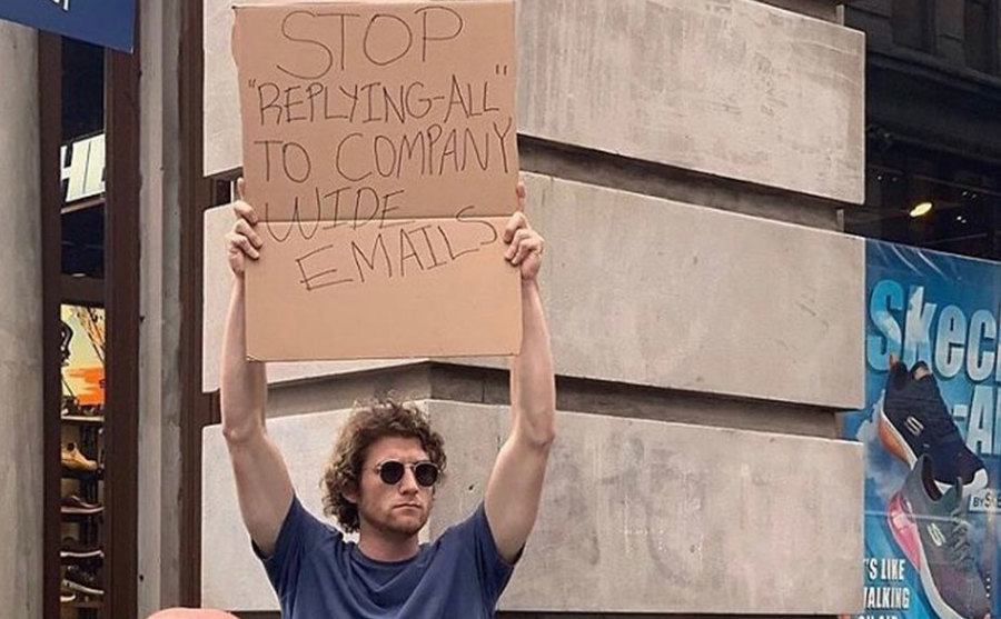 A man is holding a sign that reads ‘’ Stop Replying – All to Company-Wide Emails.’’