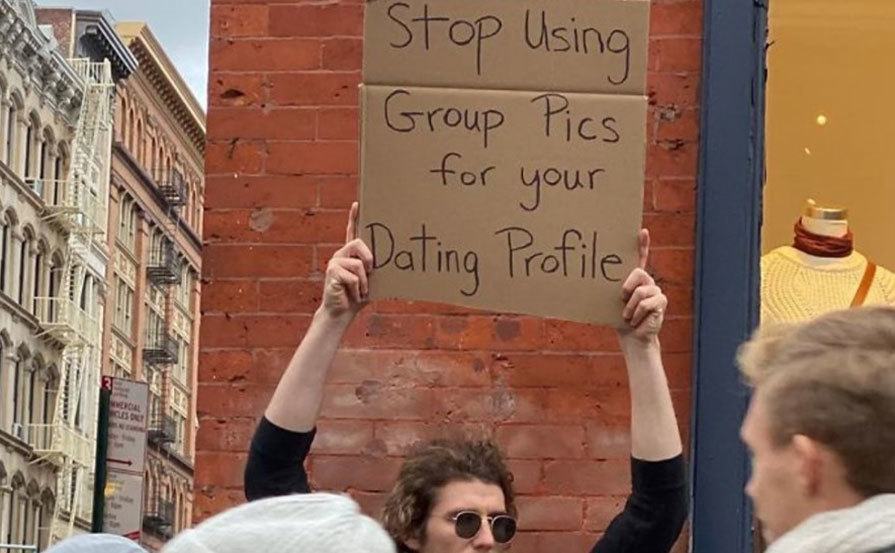 A man is holding a sign that reads ‘’Stop Using Group Pics for your Dating Profile.’’