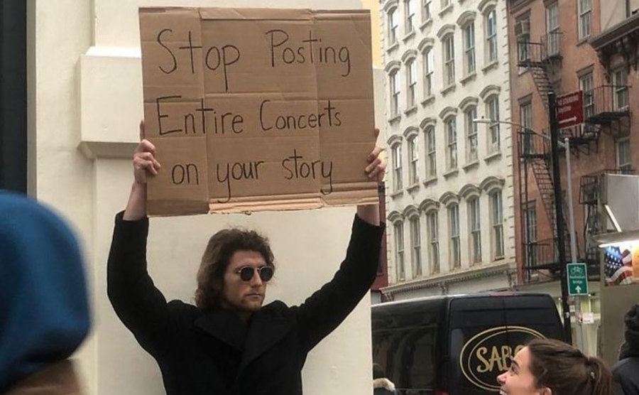 A man is holding a sign that reads ‘’Stop Posting Entire Concerts on Your Story.’’