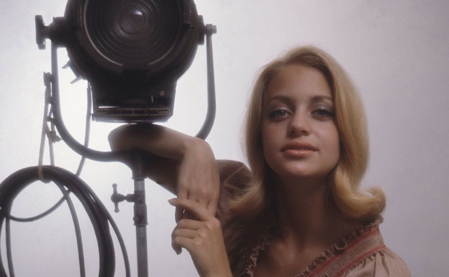 Goldie Hawn, in a pink dress, leans on a studio light.