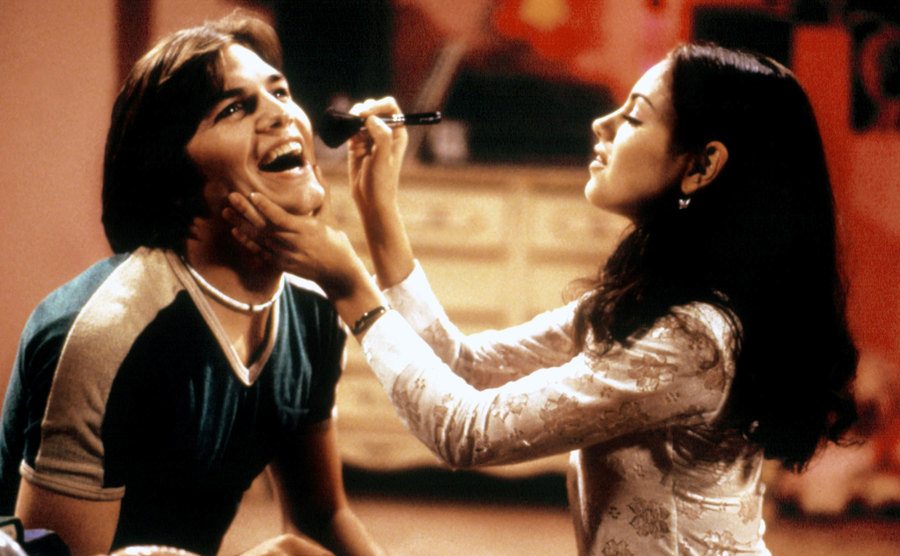 Kunis is applying makeup to Kutcher’s face in a still from That 70’s Show.