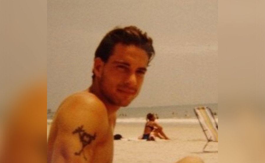 Brian sits on the beach; his tattoo is visible on his arm.