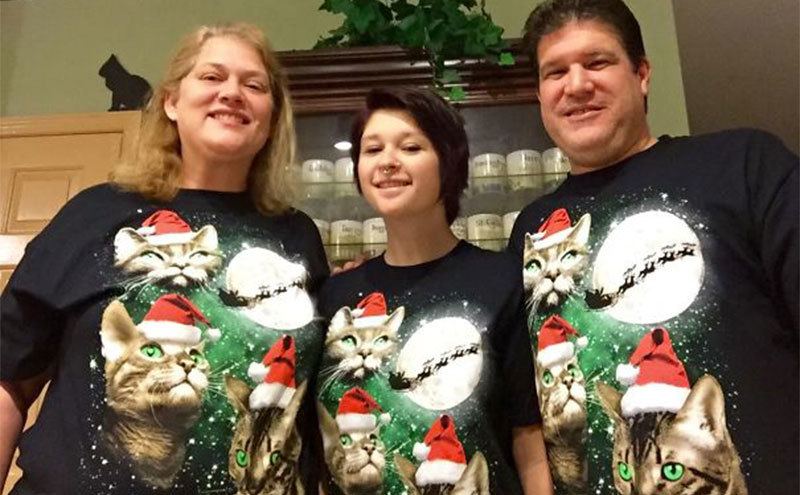 Three people are wearing matching sweaters with cats and a Santa slay on them. 