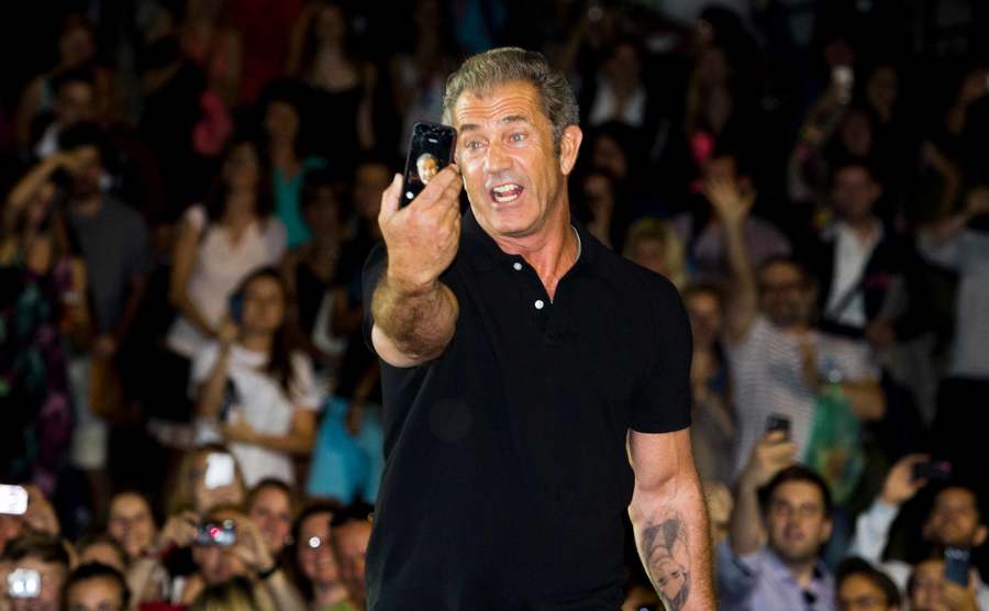 Mel Gibson takes a selfie as he stands on stage in front of a crowd.