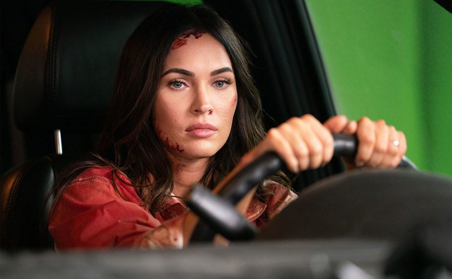 Fox sits in the driver's seat of a car on a green screen stage.