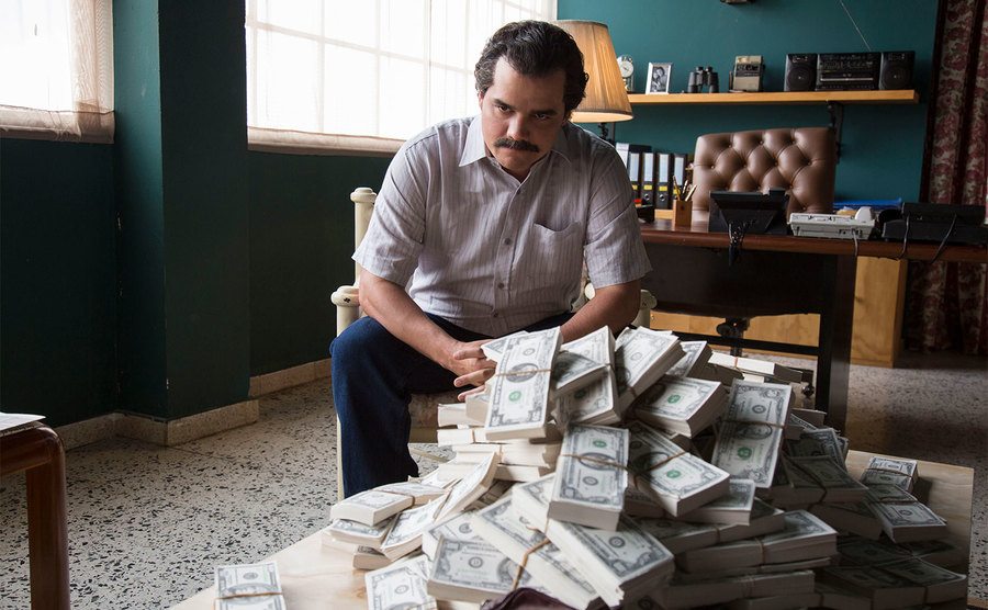Wagner Moura, AS Pablo Escobar, sits in front of a pile of cash.