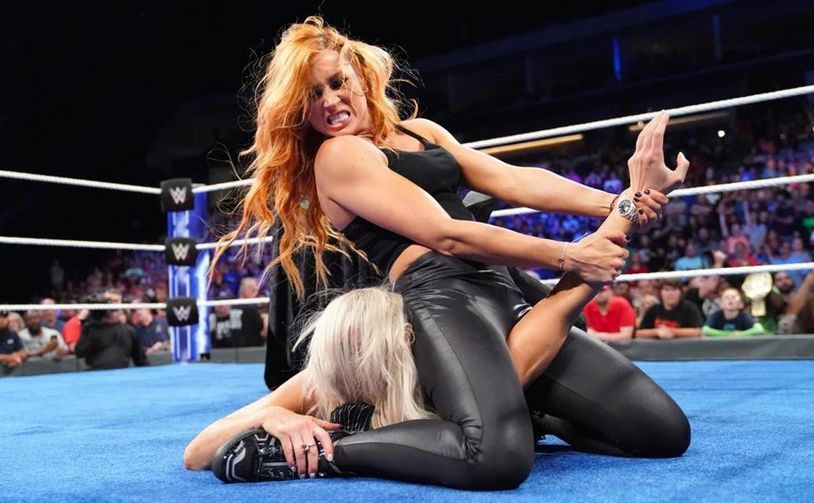 Becky sits on top of another wrestler as she is pinned under Becky's legs.