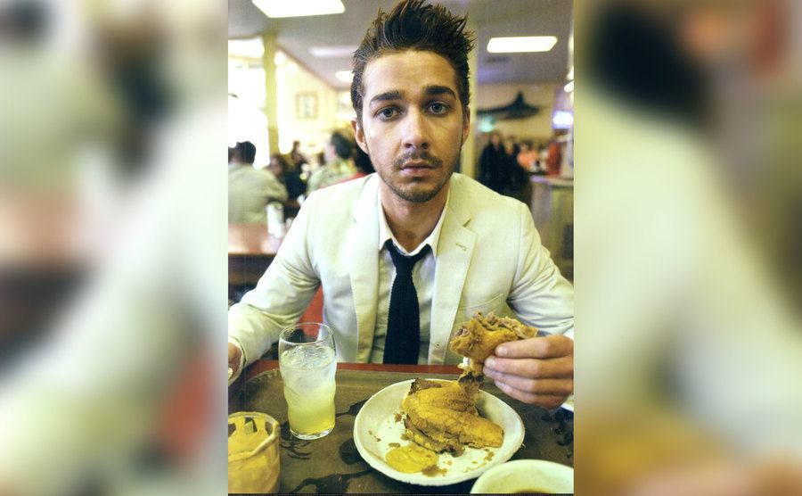 Shia sits in a restaurant eating a burger. 