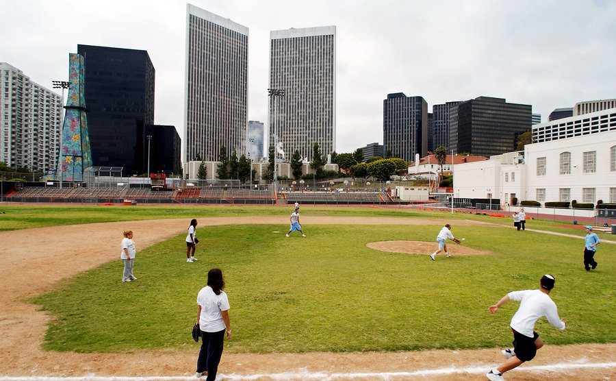 An image of high school students playing softball within sight of an oil well tower covered in flower designs.