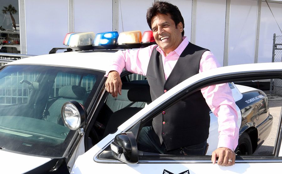 Erik Estrada poses next to a police car.