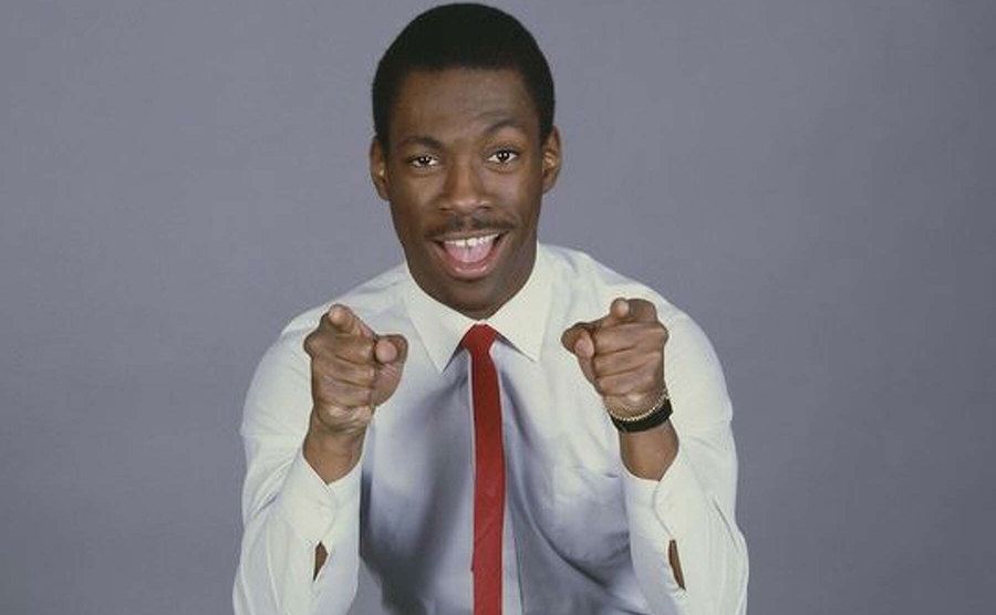 A portrait of Eddie Murphy, dressed in jeans, a white shirt, and a red necktie, as he points his fingers at the camera. 