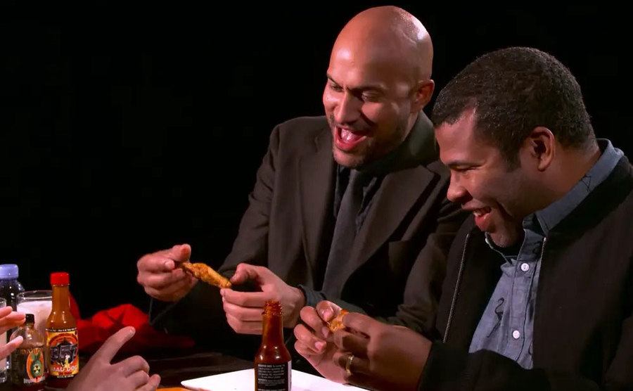 Keegan-Michael Key and Jordan Peele laugh, biting into a hot wing. 