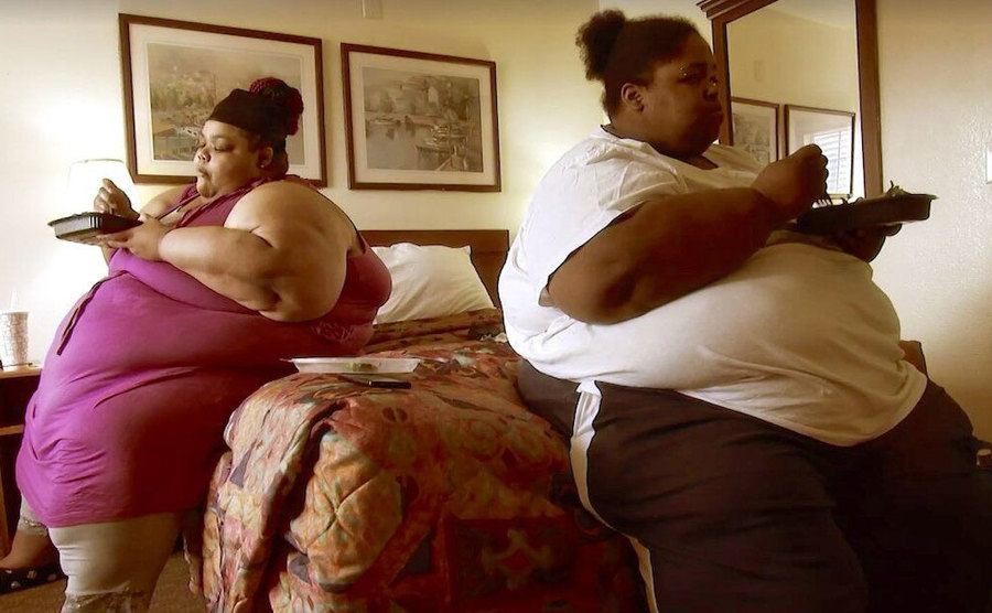 Two overweight women are sitting in the bedroom eating.